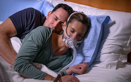 A woman, wearing a ResMed AirFit F40 mask, sleeps on her side in bed next to her husband.